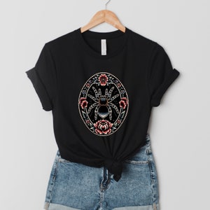 May include: Black t-shirt with a graphic of a spider surrounded by pink and red flowers in a circular design.