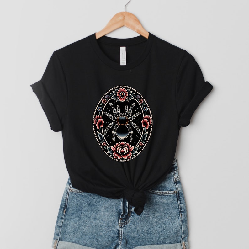 May include: Black t-shirt with a graphic of a spider surrounded by pink and red flowers in a circular design.