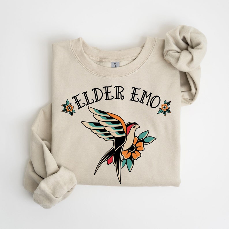 May include: A beige sweatshirt with the text "ELDER EMO" printed in black. A stylized tattoo-style bird with colorful wings and a flower is printed below the text.