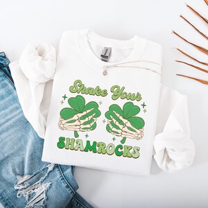 Puede incluir: Sudadera blanca con un dise&ntilde;o de tr&eacute;bol verde y el texto "Shake Your Shamrocks".