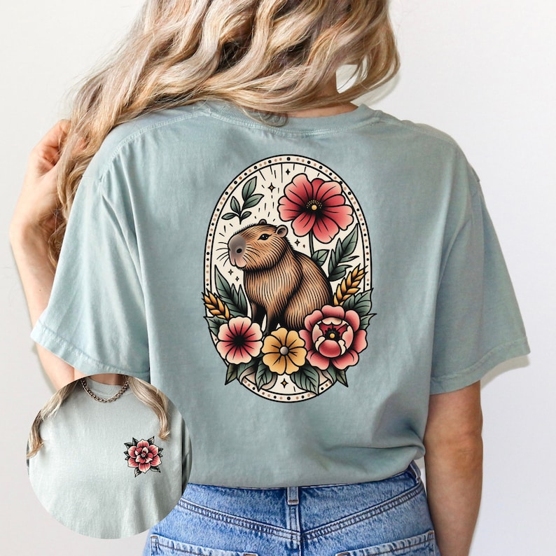 K&ouml;nnte beinhalten: Ein hellblaues T-Shirt mit einer Vintage-Illustration eines Capybaras, umgeben von Blumen. Die Illustration befindet sich in einem ovalen Rahmen mit einem floralen Rand.