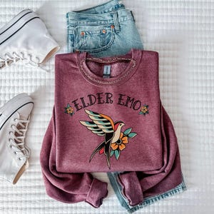 May include: A maroon sweatshirt with the text "ELDER EMO" and a vintage-style tattoo of a bird with flowers.