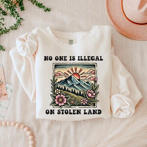 May include: A white sweatshirt with a graphic print of a mountain landscape with a rising sun and the text "No one is illegal on stolen land."