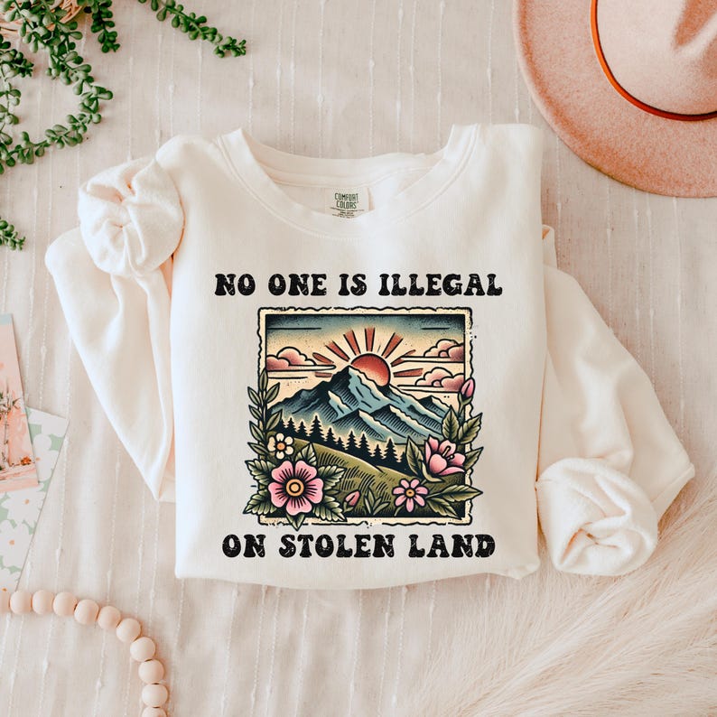 May include: A white sweatshirt with a graphic print of a mountain landscape with a rising sun and the text "No one is illegal on stolen land."