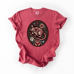 K&ouml;nnte beinhalten: Ein rotes T-Shirt mit einem floralen Design, in dessen Mitte sich eine Ratte befindet. Die Ratte ist braun und grau mit einem langen Schwanz. Die Blumen sind rosa, rot und wei&szlig;.