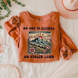 May include: A rust-colored sweatshirt with the text "NO ONE IS ILLEGAL ON STOLEN LAND" in black. The text is surrounded by a vintage-style illustration of a mountain range with a sunset and flowers.