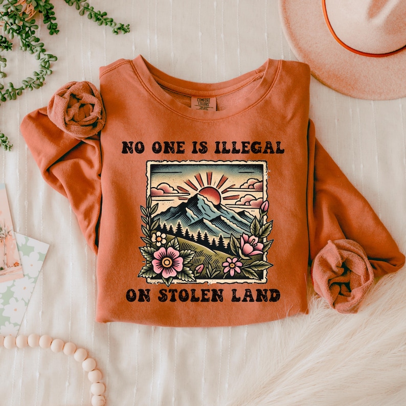 May include: A rust-colored sweatshirt with the text "NO ONE IS ILLEGAL ON STOLEN LAND" in black. The text is surrounded by a vintage-style illustration of a mountain range with a sunset and flowers.