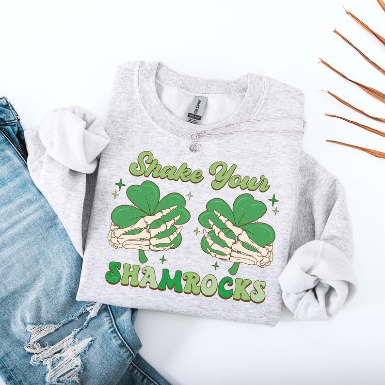 Puede incluir: Una sudadera gris con el texto "Shake Your Shamrocks" en una fuente retro. El texto est&aacute; rodeado de dos tr&eacute;boles verdes sostenidos por manos de esqueleto.