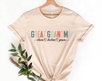 May include: A beige t-shirt with the text "GREAT GRANDMA" in colorful letters. Below the text, it says "olivia ♥ declan ♥ jason" in black script.