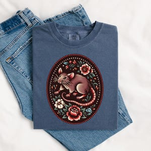 K&ouml;nnte beinhalten: Ein marineblaues T-Shirt mit einem floralen Design, das eine braune Ratte zeigt. Die Ratte ist von roten, wei&szlig;en und rosa Blumen umgeben.