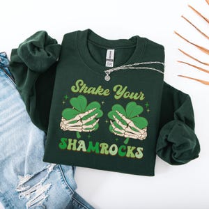 Puede incluir: Una sudadera verde oscuro con el texto "Shake Your Shamrocks" en una fuente retro. El gr&aacute;fico presenta dos manos esquel&eacute;ticas sosteniendo tr&eacute;boles.