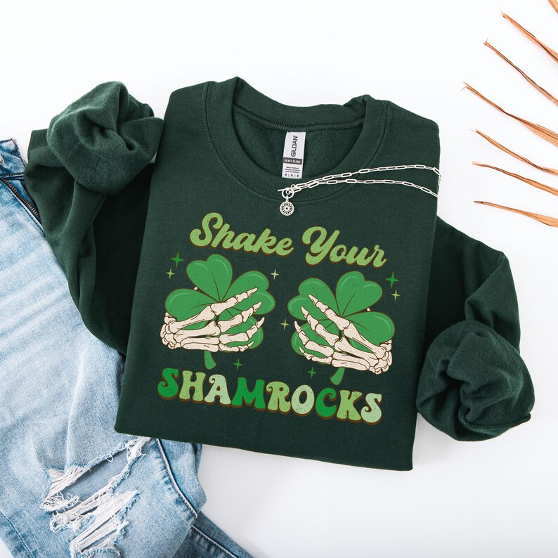 Puede incluir: Una sudadera verde oscuro con el texto "Shake Your Shamrocks" en una fuente retro. El gr&aacute;fico presenta dos manos esquel&eacute;ticas sosteniendo tr&eacute;boles.