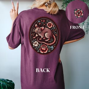 May include: A maroon t-shirt with a floral design featuring a rat in the center. The design is surrounded by flowers and leaves in shades of red, pink, and green. The back of the shirt says "BACK" in white text.