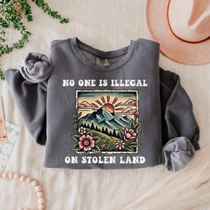 May include: A dark gray sweatshirt with a white graphic print of a mountain landscape with a rising sun and the text "NO ONE IS ILLEGAL ON STOLEN LAND".