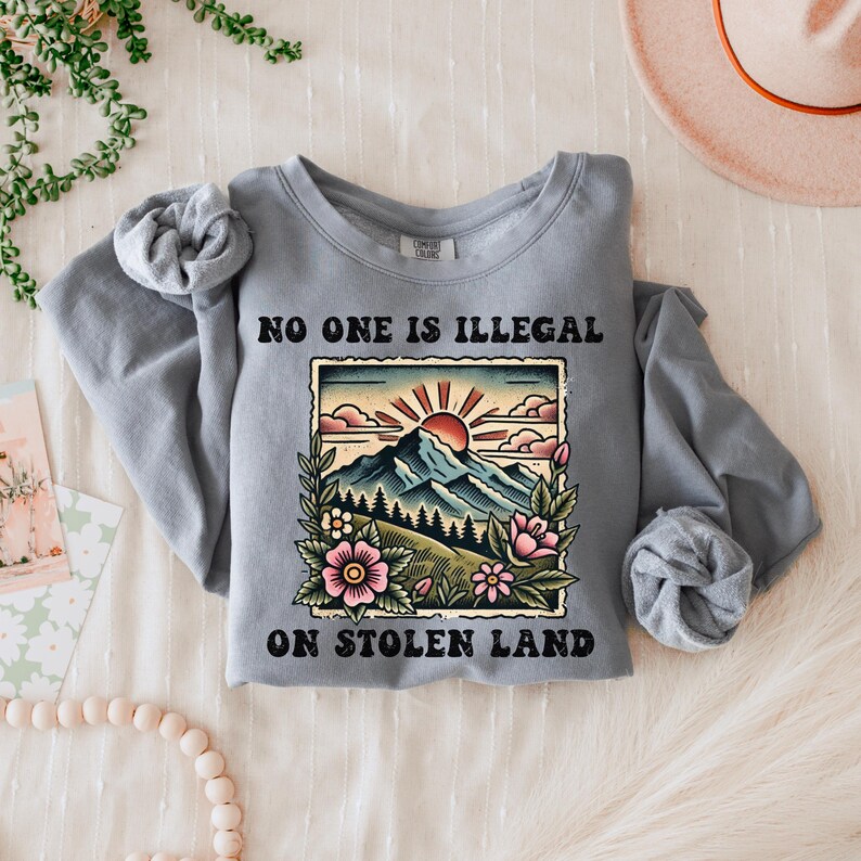 May include: Gray sweatshirt with a graphic of a mountain landscape with a sun and flowers. The text "No one is illegal on stolen land" is printed on the sweatshirt.