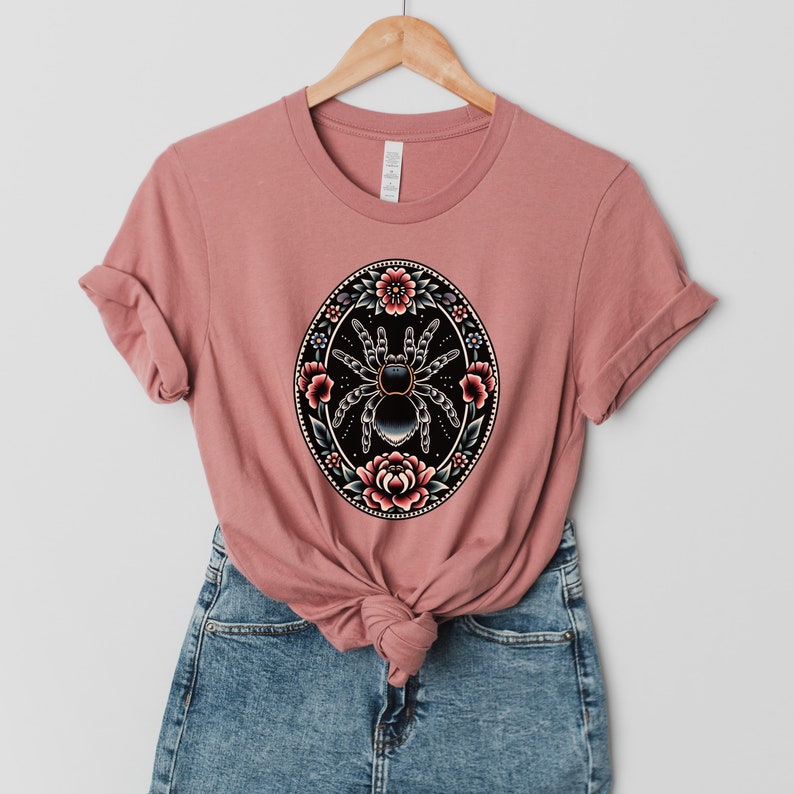 May include: A dusty rose t-shirt with a black and white spider surrounded by pink and blue flowers in a circular design.