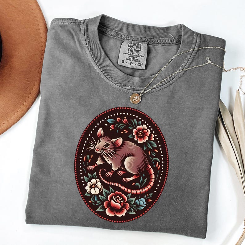 K&ouml;nnte beinhalten: Ein graues T-Shirt mit einer Vintage-Illustration einer Ratte, die von Blumen umgeben ist. Die Ratte ist braun und wei&szlig;, die Blumen sind rot, rosa und blau. Das Shirt hat den Text "COMFORT COLORS" und "S - P - CH" auf der Vorderseite.
