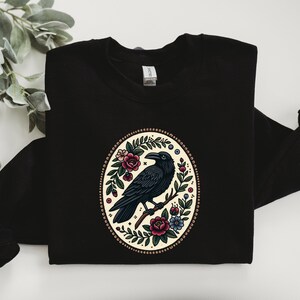 May include: Black sweatshirt with a circular design featuring a black raven perched on a branch surrounded by colorful flowers and leaves. The design is on a cream background with a brown beaded border.