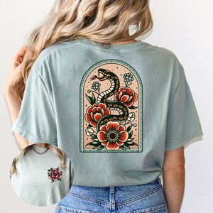 K&ouml;nnte beinhalten: Ein hellblaues T-Shirt mit einem Tattoo-Design im Vintage-Stil auf der R&uuml;ckseite. Das Design zeigt eine schwarze Schlange, die sich um rote und wei&szlig;e Blumen in einem pfirsichfarbenen Bogen windet.