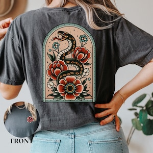 Pode incluir: Uma camiseta preta com um design de tatuagem vintage de uma cobra enrolada em flores em um arco cor de pêssego. O design está na parte de trás da camiseta. A parte da frente da camiseta tem um pequeno design de flor.