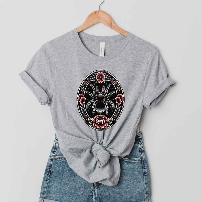 May include: A gray t-shirt with a black and white spider surrounded by pink and red flowers in a circular design.