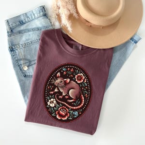 K&ouml;nnte beinhalten: Ein maroonfarbenes T-Shirt mit einem Aufdruck eines braunen Ratten, umgeben von bunten Blumen in einem runden Rahmen.