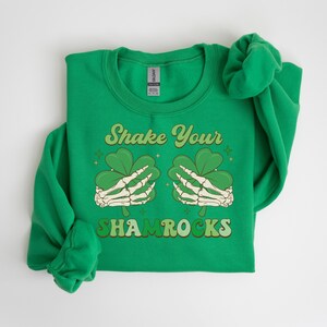 Puede incluir: Sudadera verde con el texto "Shake Your Shamrocks" en una fuente retro. El gr&aacute;fico presenta dos manos esquel&eacute;ticas sosteniendo tr&eacute;boles.