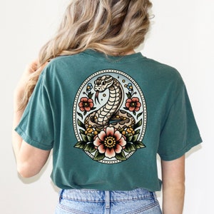 May include: A teal blue t-shirt with a vintage style tattoo design of a snake with flowers on the back.
