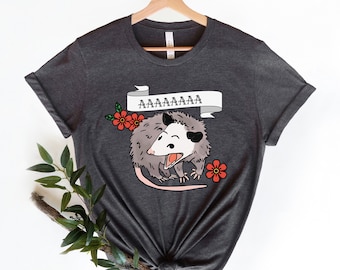 Puede incluir: Una camiseta gris oscuro con una ilustración de dibujos animados de una zarigüeya con el texto "AAAAAAA" encima. La zarigüeya es blanca con ojos negros y una nariz y boca rosadas. Está rodeada de flores rojas.