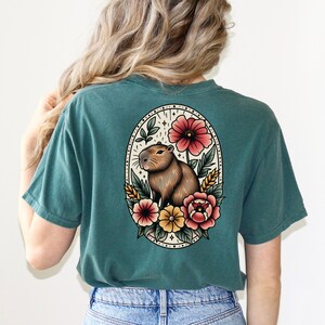 K&ouml;nnte beinhalten: Ein blaugr&uuml;nes T-Shirt mit einer Vintage-Illustration eines Capybaras, umgeben von Blumen. Die Illustration befindet sich in einem runden Rahmen mit schwarzem Rand.
