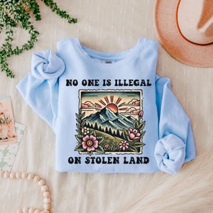 May include: Light blue sweatshirt with the text "No one is illegal on stolen land" in black lettering. A vintage-style illustration of a mountain range with a sun and flowers is above the text.