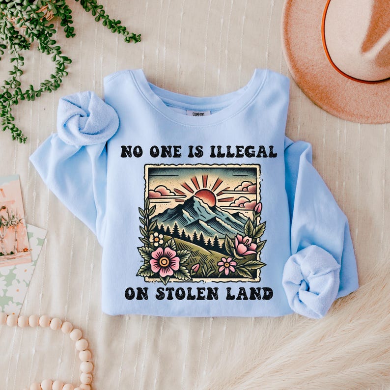 May include: Light blue sweatshirt with the text "No one is illegal on stolen land" in black lettering. A vintage-style illustration of a mountain range with a sun and flowers is above the text.