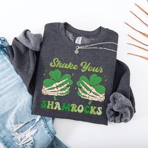 Puede incluir: Una sudadera gris oscura con el texto "Shake Your Shamrocks" en una fuente retro. El gr&aacute;fico presenta dos manos esquel&eacute;ticas sosteniendo tr&eacute;boles.