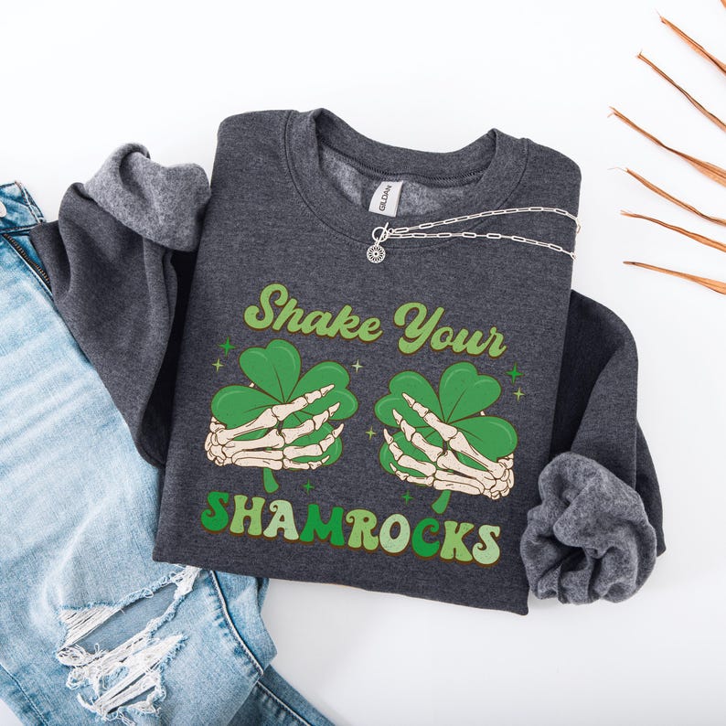 Puede incluir: Una sudadera gris oscura con el texto "Shake Your Shamrocks" en una fuente retro. El gr&aacute;fico presenta dos manos esquel&eacute;ticas sosteniendo tr&eacute;boles.