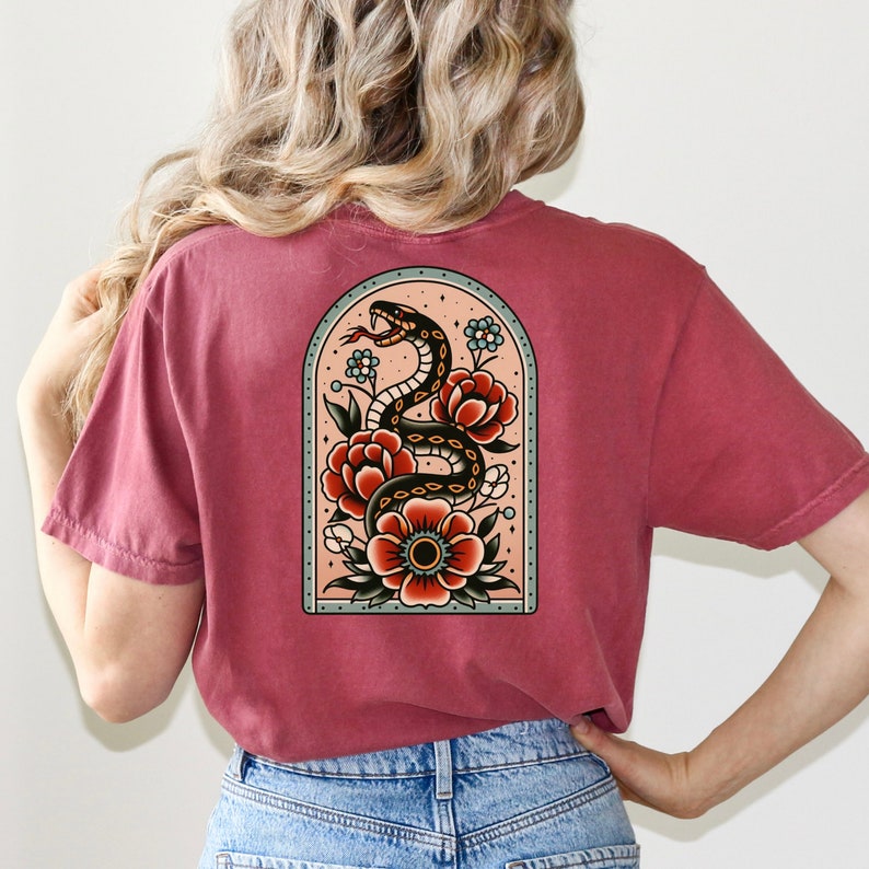 K&ouml;nnte beinhalten: Ein maronfarbenes T-Shirt mit einem Tattoo-Design im Vintage-Stil, das eine schwarze Schlange zeigt, die sich um rote und wei&szlig;e Blumen in einem blauen und gr&uuml;nen Bogen windet.