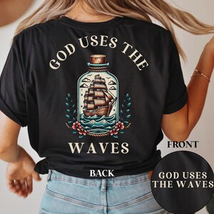 Könnte beinhalten: Schwarzes T-Shirt mit einem Grafikdesign eines Schiffs in einer Flasche, umgeben von Blumen und Blättern. Der Text "GOD USES THE WAVES" ist über und unter dem Bild angeordnet. Das Wort "BACK" ist darunter.