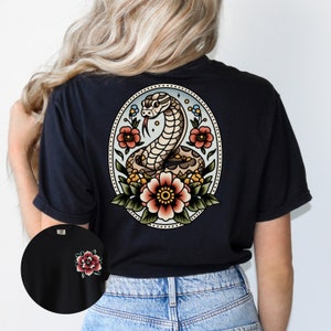 May include: Black t-shirt with a vintage-style tattoo design of a snake with flowers on the back. The design is in an oval frame with a blue background.