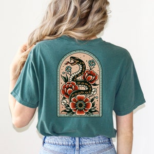 K&ouml;nnte beinhalten: Ein blaues T-Shirt mit einem Tattoo-Design im Vintage-Stil, das eine schwarze Schlange zeigt, die sich um rote und orangefarbene Blumen windet. Das Design befindet sich in einem gew&ouml;lbten Rahmen mit einem hellrosa Hintergrund.