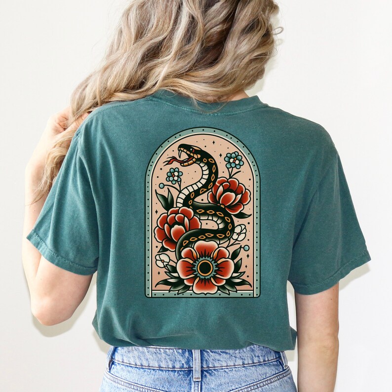 K&ouml;nnte beinhalten: Ein blaues T-Shirt mit einem Tattoo-Design im Vintage-Stil, das eine schwarze Schlange zeigt, die sich um rote und orangefarbene Blumen windet. Das Design befindet sich in einem gew&ouml;lbten Rahmen mit einem hellrosa Hintergrund.