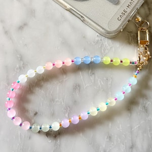 May include: A clear phone case with a glittery back and a silver magnetic ring. The case is attached to a beaded wrist strap with pastel-colored beads. The strap has a gold-colored clasp and the words "CASE-MATE" are printed on the case.