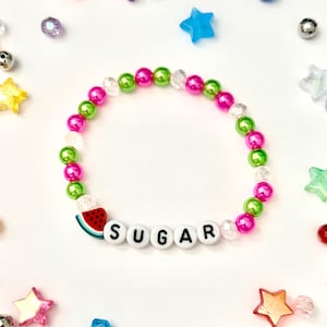 May include: A beaded bracelet with pink, green, and clear beads. The bracelet features the word "SUGAR" spelled out in white letter beads and a watermelon charm. Scattered around the bracelet are colorful star and round beads.