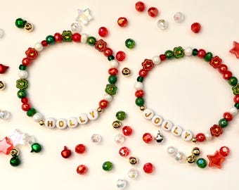 May include: Two beaded bracelets with the words "HOLLY" and "JOLLY" spelled out in white letter beads. The bracelets feature red, green, white, and gold beads, with flower-shaped accents. Scattered around are additional beads and star-shaped embellishments.