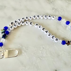 May include: A beaded lanyard with the words "PATHOLOGICAL PEOPLE PLEASER" spelled out in white letter beads. The lanyard features blue and clear beads, along with star-shaped charms and a silver clasp.