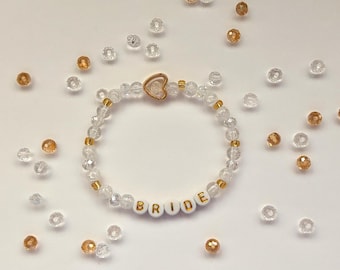 May include: A white beaded bracelet with a gold heart charm and the word "BRIDE" spelled out in gold letters on white beads.