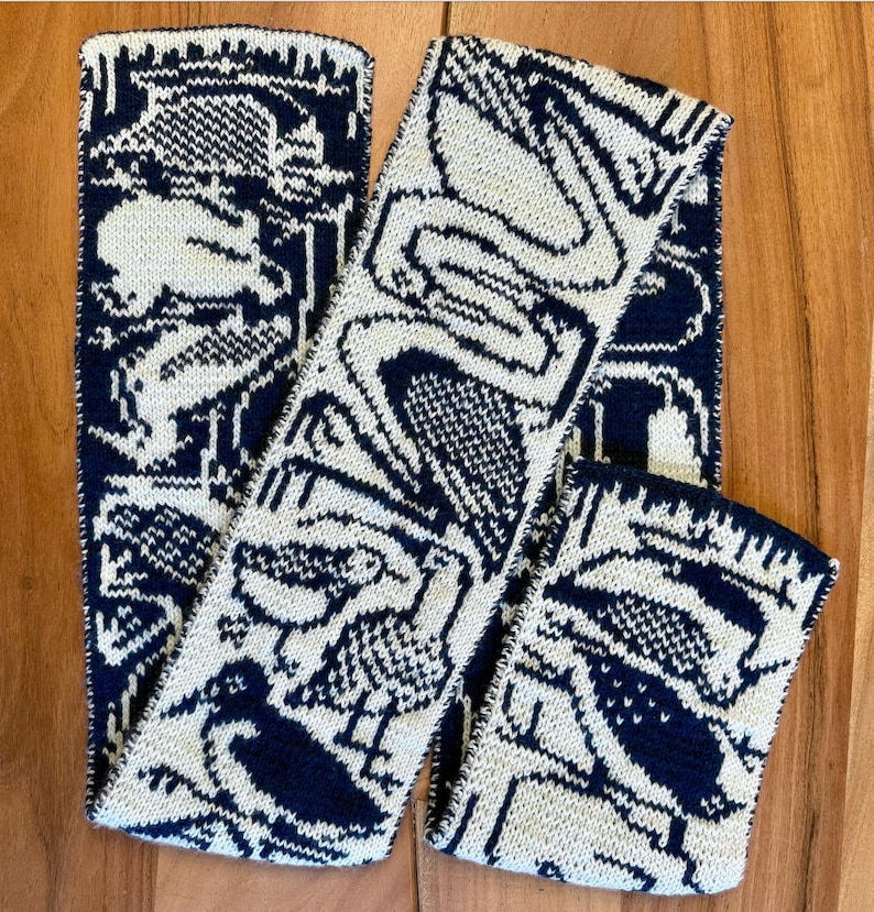 Puede incluir: Bufanda de punto y calentadores de mu&ntilde;eca a juego en crema y azul marino. El patr&oacute;n presenta aves estilizadas y formas geom&eacute;tricas. La bufanda est&aacute; doblada y los calentadores de mu&ntilde;eca est&aacute;n sobre una superficie de madera.