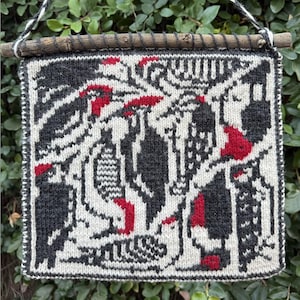 May include: A square, hand-knitted wall hanging featuring a black, white, and red bird design. The artwork is mounted on a small wooden dowel with a twisted cord for hanging. The background is a lush green foliage.
