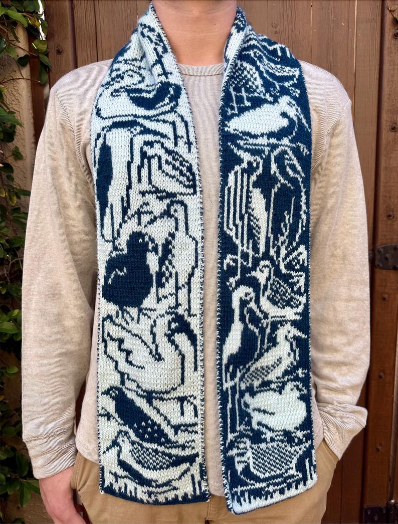 Puede incluir: Bufanda de punto con un patr&oacute;n repetido de aves en azul marino y blanco roto. La bufanda est&aacute; drapeada alrededor del cuello, mostrando el dise&ntilde;o detallado. La bufanda est&aacute; hecha de un material suave.