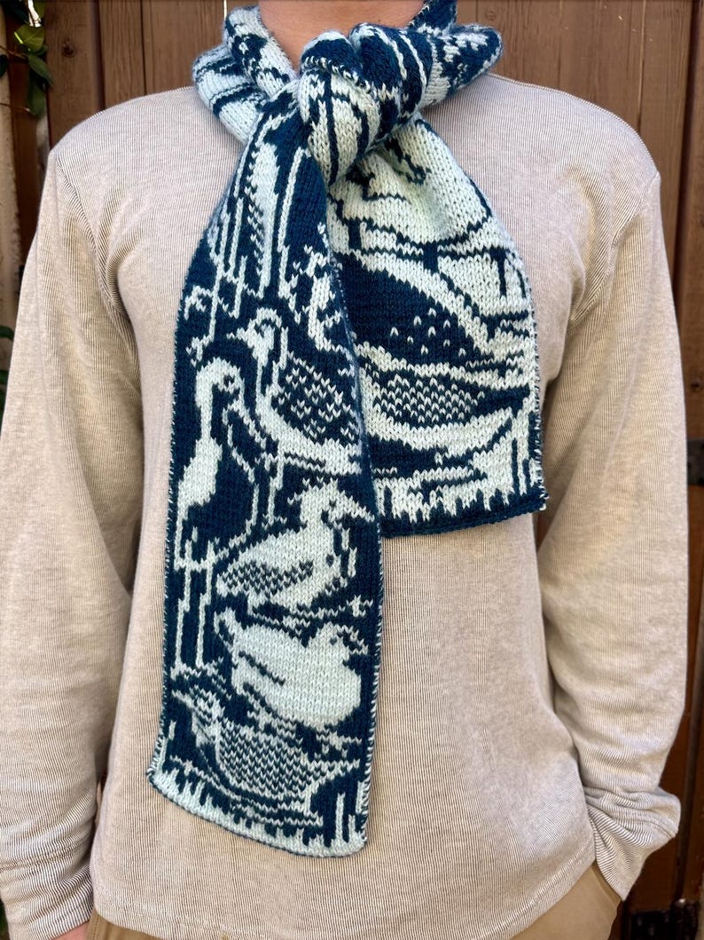 Puede incluir: Bufanda de punto con un patr&oacute;n azul marino y blanco roto que presenta aves y formas abstractas. La bufanda est&aacute; envuelta alrededor del cuello, mostrando el dise&ntilde;o detallado y la textura del tejido. La bufanda est&aacute; hecha de material suave.