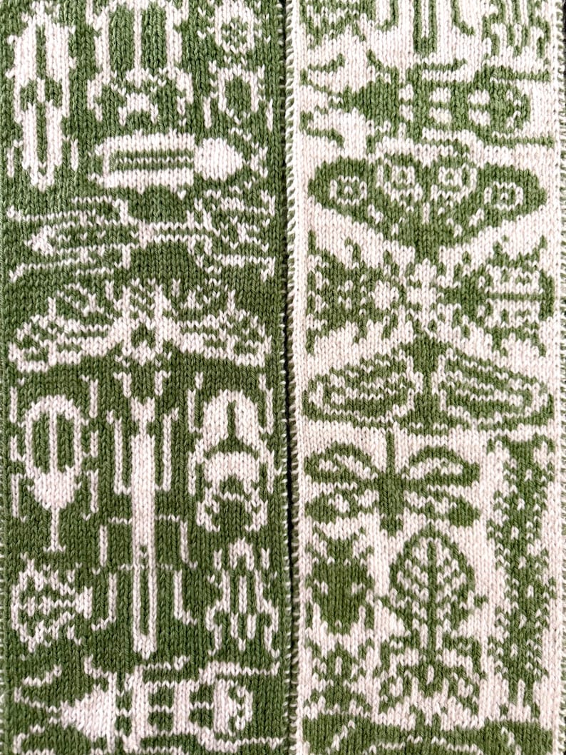 Op de afbeelding: Close-up van een gebreid textiel met een herhalend patroon in groen en wit. Het ontwerp bevat gestileerde vormen en symbolen, waardoor een gestructureerd, decoratief oppervlak ontstaat. De stof lijkt een sjaal of een vergelijkbaar item te zijn.