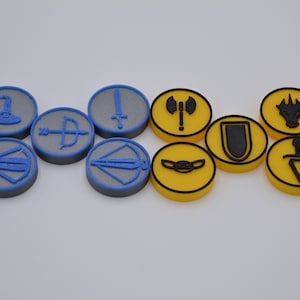 May include: A set of ten round tokens with different symbols. Five tokens are gray with blue trim and five tokens are yellow with black trim. The symbols include a sword, an axe, a shield, a dragon, a bow and arrow, a wizard's hat, and a dagger.